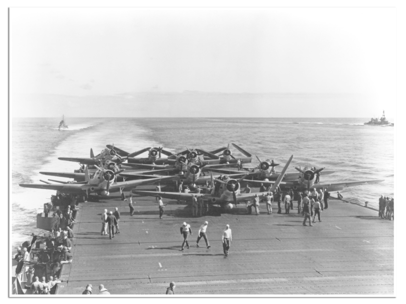 een vliegdek vol propellervliegtuigen met op de voorgrond personeel