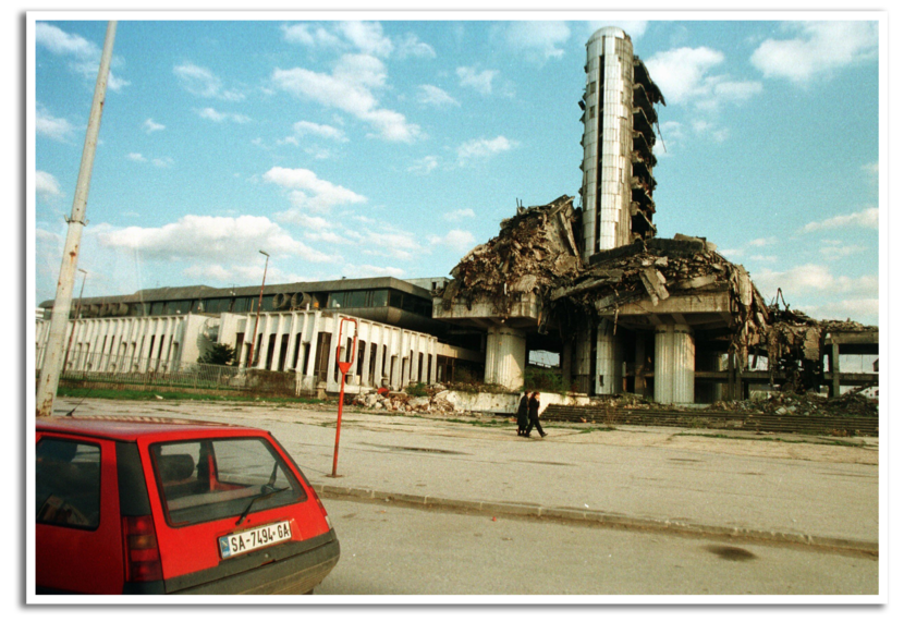Een rode auto staat geparkeerd voor het verwoeste gebouw van de tv-zender van Sarajevo.
