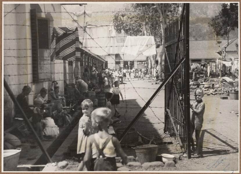 Op de voorgrond een omheining van prikkeldraad met rechts van het midden een geopend hek. Voor het hek en voor de omheining staan kinderen. Op de achtergrond een deel van het kamp met op de stoepen voor de gebouwen veel vrouwen en kinderen.