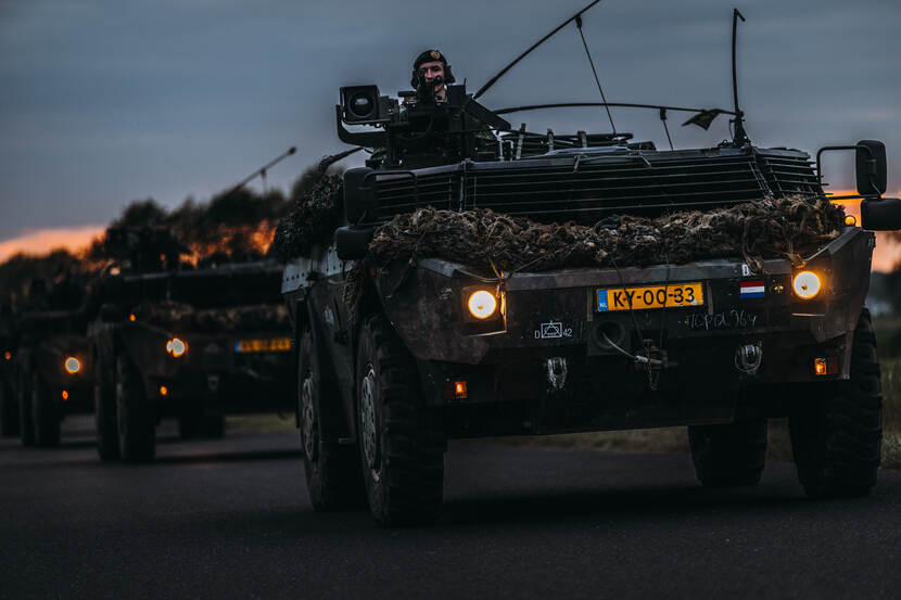 Een Fennek verkenningsvoertuig rijdt over de weg met achter zich de ondergaande zon.