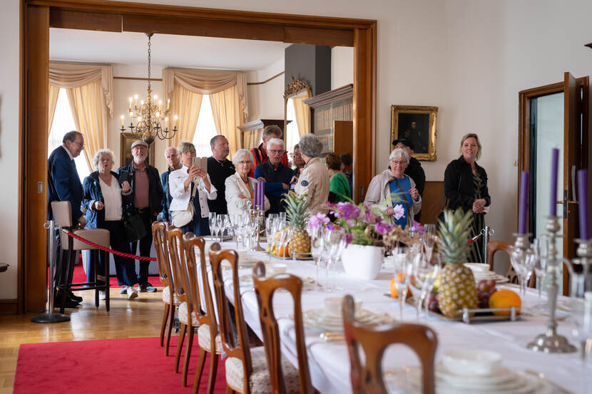 Lange gedekte tafel met wit linnen en porseleinen servies waarlangs belangstellenden lopen.