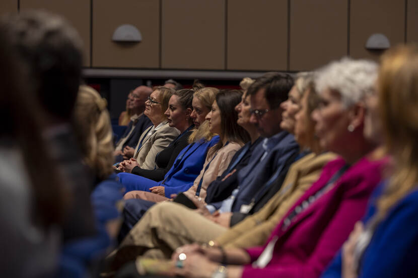 Een aantal personen zit naast elkaar en luistert aandachtig naar de spreker op het podium.