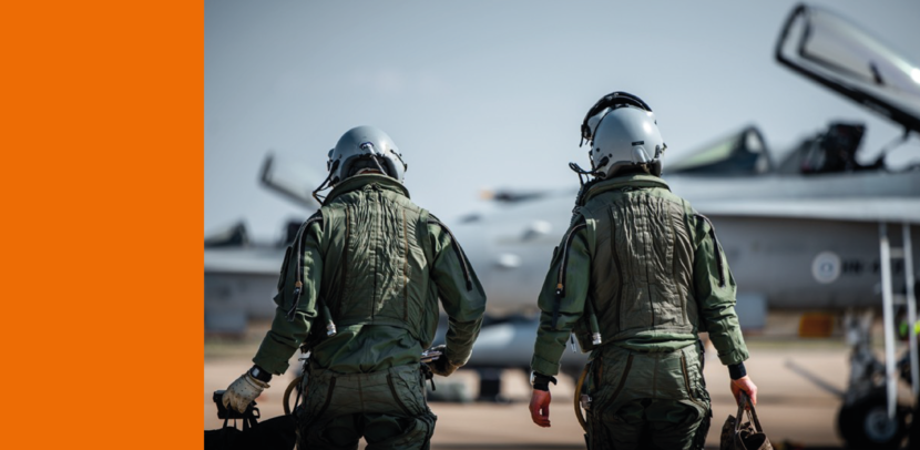 Twee vliegers, van de achterzijde bezien, dragen een helm en lopen langs jachtvliegtuigen. Die zijn vaak op de achtergrond te zien.