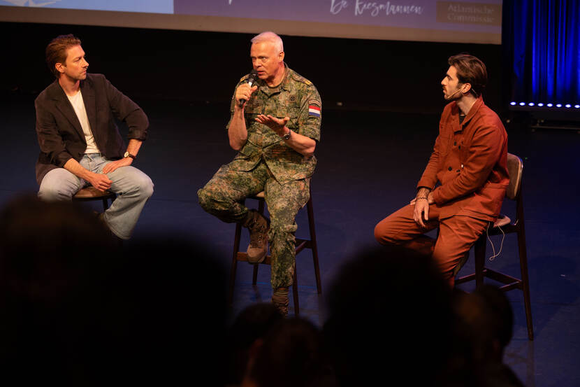 Militair met microfoon in de hand zit op barkruk op het podium van een theater. Links en rechts van hem zitten twee mannen in burgerkleding op een barkruk.