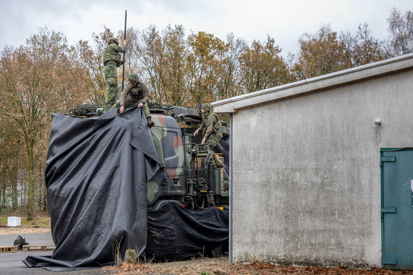Militairen camoufleren een vrachtwagencabine met zwart zeildoek. De truck staat naast een grijs gebouw.