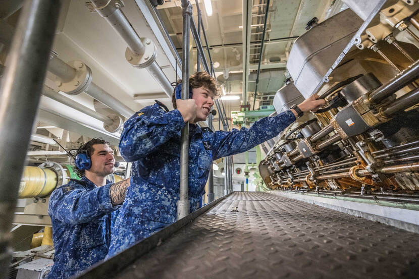 Een lachende matroos werkt in de technische ruimte van het schip, terwijl een collega hem ondersteunt.