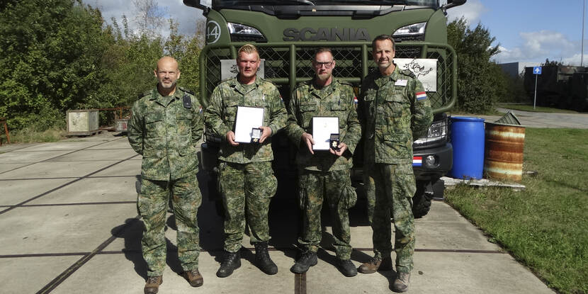 Sergeanten der eerste klasse Bastiaan Holst en Robbin van den Berg met hun waarderingsspelden te midden van Commandant Defensie Helikopter Commando en Commandant 930 Squadron voor een Scania-vrachtwagen.
