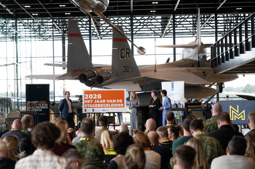 Tuinman spreekt voor publiek in het Nationaal Militair Museum. Naast hem een bord met de tekst ‘2026; het jaar van de stagebegeleider’, achter hem een tentoongesteld vliegtuig.