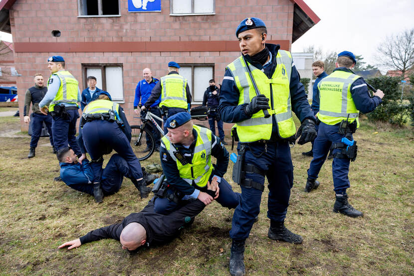 Twee marechaussees werken ieder een verdachte naar de grond. Vier andere collega’s vormen een cirkel rondom de aanhoudingen en houden daarmee de omstanders op afstand.
