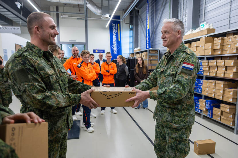 Een man in uniform draagt een doos over aan een andere man in uniform, op de achtergrond mannen met een oranje trainingspak.