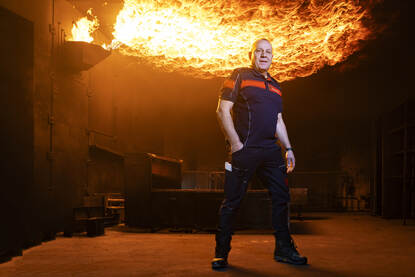 Een man staat in een stoere houding in een donkere ruimte met op de achtergrond een enorme steekvlam die met een computer aan- en uitgezet kan worden.