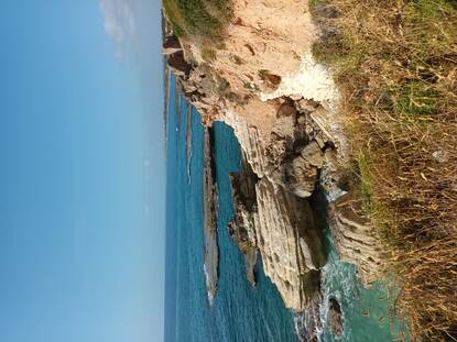 Een foto van een groen begroeide klif met op de achtergrond een azuurblauwe hemel en water.