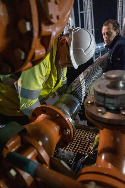 Een collega van de marine kijkt mee over de schouder van een Engelsman terwijl hij de tankslang koppelt aan het schip.