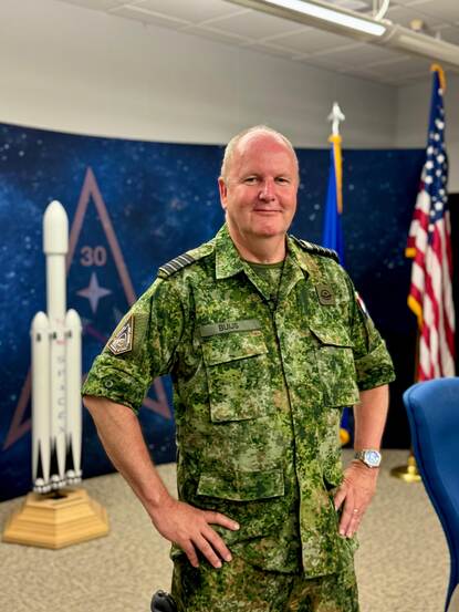 Portretfoto van kolonel Bernard Buijs, achter hem een Amerikaanse vlag en een modelversie van een raket.