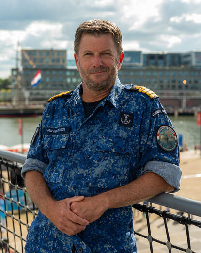 Een marineman poseert voor een hek met op de achtergrond een zeilschip en gebouwen.