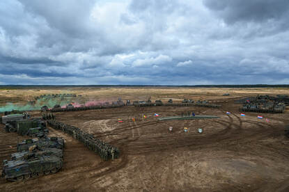 Links: Voertuigen en militairen van de battlegroup staan op een veld, met in het midden de vlaggen van de deelnemers. Rechts twee militaire pantservoertuigen rijden door rook en stof op een heuvelachtig terrein, omringd door bos.