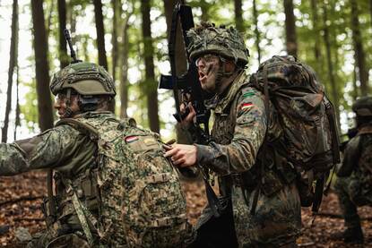 twee militairen in camouflagetenue liggen achter een lage muur, gericht op de verte met een groot geweer op een statief, omringd door bos en open veld. Rechts: Twee soldaten in camouflagetenue staan in een bos; één helpt de ander met zijn rugzak, terwijl de ander een geweer vasthoudt en iets roept.