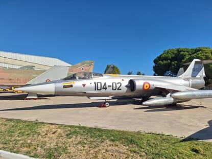 links en rechts een militair vliegtuig dat buiten op een grasveld staat. Ze staan op een droog, dor grasveld met enkele bomen en struiken op de achtergrond. De lucht is helderblauw en het is duidelijk een zonnige dag.