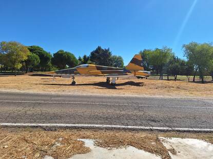 links en rechts een militair vliegtuig dat buiten op een grasveld staat. Ze staan op een droog, dor grasveld met enkele bomen en struiken op de achtergrond. De lucht is helderblauw en het is duidelijk een zonnige dag.