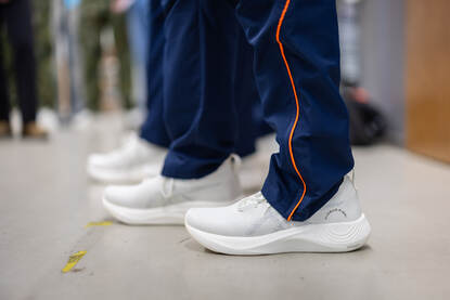 Twee personen dragen witte, moderne sportschoenen met een dikke, gebogen zool en een donkerblauwe broek met oranje strepen langs de zijkant. Rechts: Twee personen in camouflagebroek dragen witte, moderne sportschoenen met dikke zolen; een persoon bindt de veters vast,.