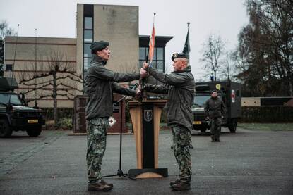 Twee militairen staan tegenover elkaar met een militaire vlag in het midden. Op de achtergrond staan een militair en twee militaire voertuigen.