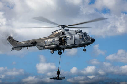 Op de linkerfoto vliegt een helikopter met eronder een soort mand, de AirTEP. Op de rechterfoto zijn operators in die mand geklommen.
