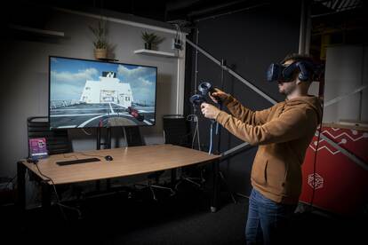 Op de foto staat een man met VR-bril op, die met in elke hand een controller voor videoscherm staat waarop een marineschip virtueel is weergegeven.