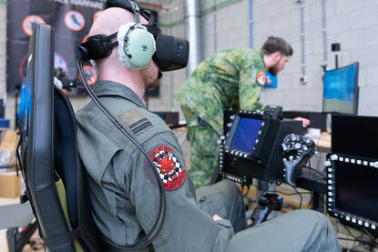 Een vlieger zit in een bureaustoel met een VR-bril op zij hoofd. Ook draagt hij een koptelefoon.