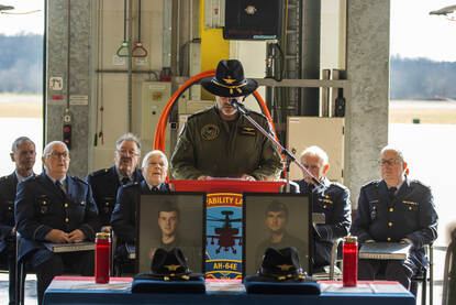 Herdenkingsbijeenkomst in hangaar, roldeuren staan open met daarachter op het platform twee Apaches.
