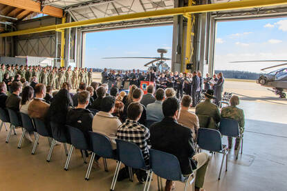 Herdenkingsbijeenkomst in hangaar, roldeuren staan open met daarachter op het platform twee Apaches.