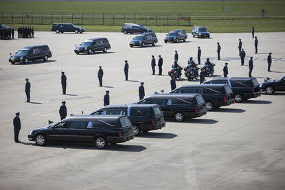 Naast een haag van luchtmachters staat een rij met rouwauto’s opgesteld. Tussen die haag rijdt een groepje motorrijders. Aan de andere kant van de haag rijden de rouwauto’s in een stoet achter elkaar, van het platform af.