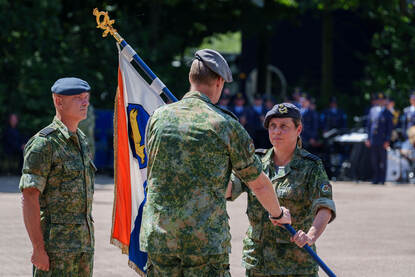 Een militair met een vlag in zijn handen staat tegenover een militair die de vlag in ontvangst gaat nemen. Een derde militair kijkt aan de zijkant toe.