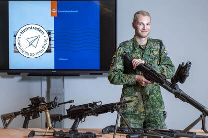 Sergeant-majoor Kees Kleinheerenbrink poseert met een paar wapens die op een tafel staan, achter hem het lesbord.
