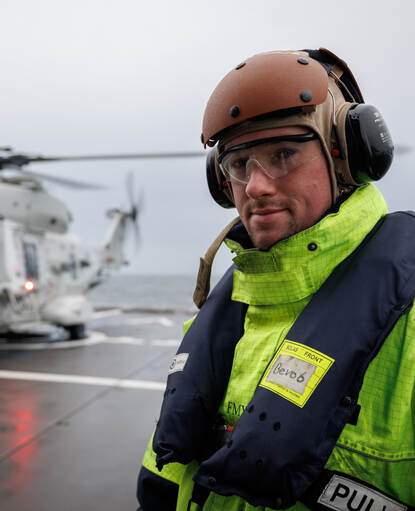 sergeant Lubbert, onderhoudsmonteur van de NH90, met gehoorbescherming en reddingsvest op het helikopterdek met op de achtergrond de NH90 boordhelikopter.