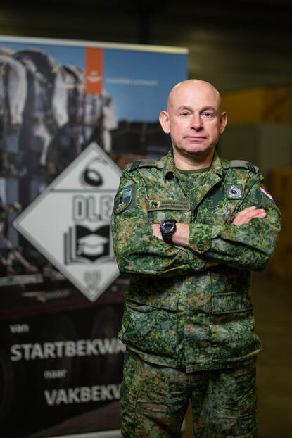 Portret van Frank Wiegman in camouflage-uniform, staand met gekruiste armen en een neutrale blik, gefotografeerd binnenshuis met op de achtergrond een informatieve banner.