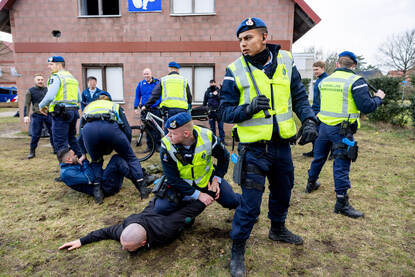 Twee marechaussees werken ieder een verdachte naar de grond. Vier andere collega’s vormen een cirkel rondom de aanhoudingen en houden daarmee de omstanders op afstand.