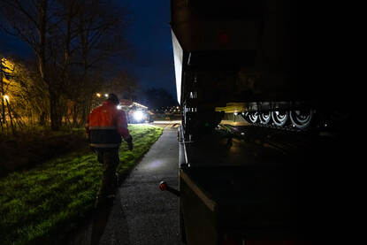 Doorkijk met rechts een pantserrupsvoertuig op een dieplader waarbij de wielen oplichten door koplampen.