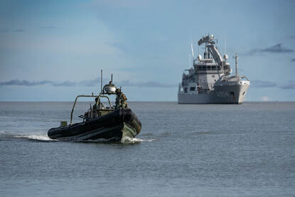 Links een Frisc-onderscheppingsvaartuig met Pelikaan op de achtergrond. Rechts mariniers in dekking met de Pelikaan op de achtergrond.