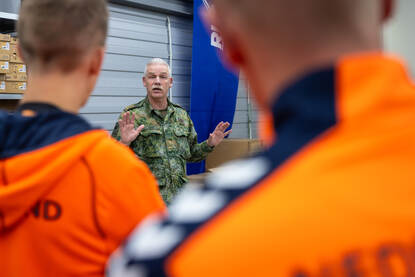 Links een man in vlekkenpak spreekt mannen in een oranje trainingsjack toe. Rechts een man die een witte schoen omhoog houdt.