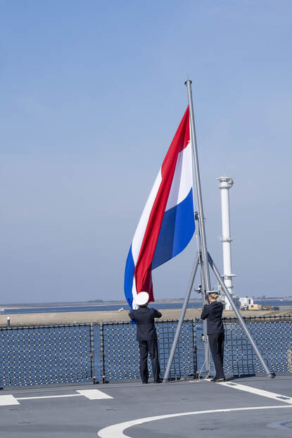 2 leden van de marine hijsen de Nederlandse vlag op het dek van een marineschip.